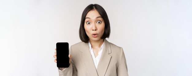 Immagine della donna aziendale asiatica che mostra lo schermo del telefono cellulare dell'interfaccia dell'app che fa l'espressione sorpresa del viso wow in piedi su sfondo bianco