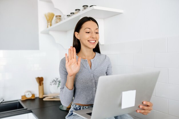 Immagine della donna bruna carina seduta sul tavolo in cucina e utilizzando il computer portatile