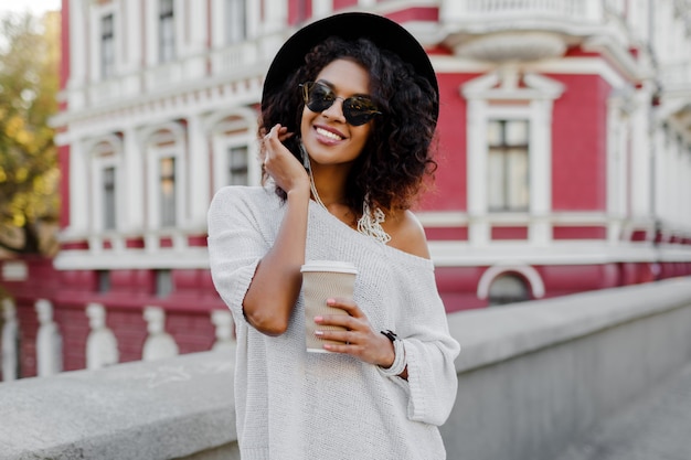 Immagine della donna di colore graziosa sorridente in maglione bianco e cappello nero che tengono tazza di caffè. Sfondo urbano.