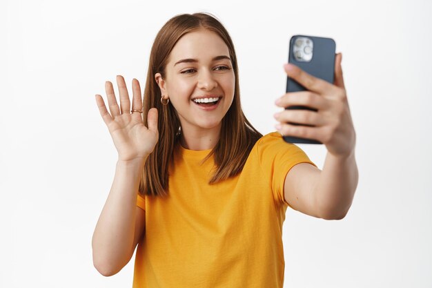 Immagine della fotocamera dello smartphone Girl Wave, videoconferenza sull'app del telefono cellulare, parlare con un amico, streaming live, sorridere e salutare, in piedi con una maglietta gialla su sfondo bianco