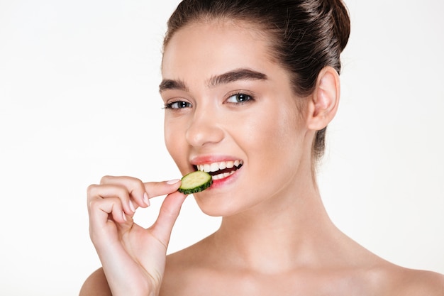 Immagine della giovane donna allegra che ha pelle pulita che mangia cetriolo che gode della disintossicazione mentre posando