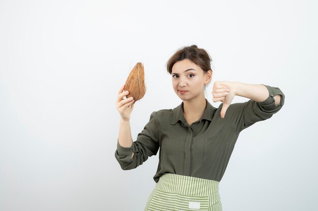 Immagine della giovane donna in grembiule che mostra il pollice giù e che tiene una noce di cocco. Foto di alta qualità