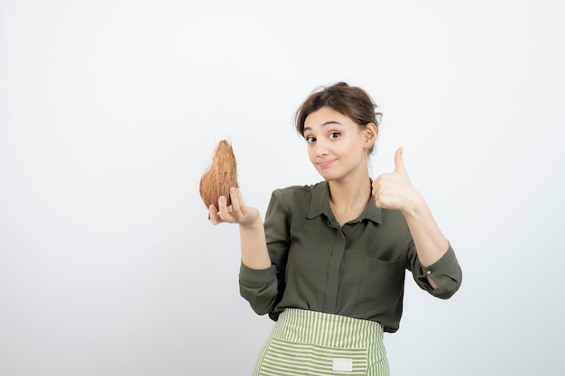 Immagine della giovane donna in grembiule che mostra il pollice in su e che tiene una noce di cocco. Foto di alta qualità