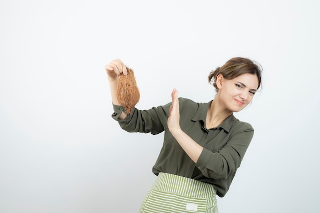 Immagine della giovane donna in grembiule che tiene una noce di cocco contro il muro bianco. Foto di alta qualità