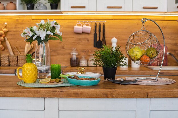 Immagine della grande cucina luminosa con armadi bianchi e marroni con bollitore per il tè all'ananas giallo, macinapepe bianco e metallo appeso con frutta e biscotti