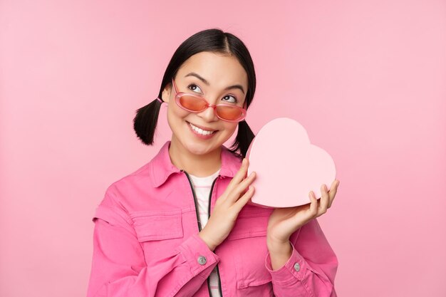 Immagine della ragazza asiatica alla moda in occhiali da sole che indovina cosa c'è all'interno della confezione regalo presente a forma di cuore in piedi su sfondo rosa