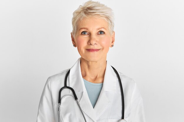 Immagine dello studio del medico femminile invecchiato centrale attraente sicuro con l'acconciatura tinta corta che posa camice bianco da portare isolato e stetoscopio.