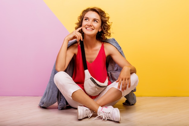 Immagine dello studio della donna adorabile castana sorridente che indossa attrezzatura sportiva alla moda e rivestimento dei jeans che si siedono sul pavimento.