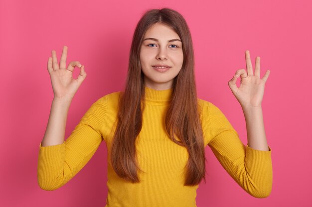 Immagine di allegro piuttosto giovane femmina alzando le braccia, mostrando il segno va bene, indossando la felpa gialla, con i capelli lunghi, essere calmo, trascorrere del tempo da solo. Concetto di emozioni.