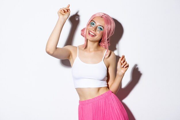 Immagine di attraente ragazza festa con parrucca rosa e trucco luminoso, divertirsi e celebrare le vacanze, ballare felice.