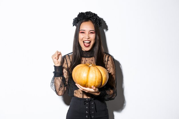 Immagine di bella donna asiatica eccitata che celebra halloween, indossando abiti da strega e tenendo la zucca, urlando di gioia.