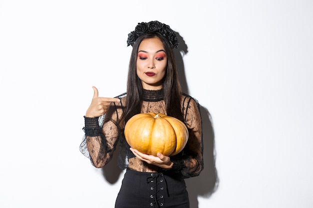 Immagine di bella donna asiatica in abito di pizzo gotico e corona nera che punta il dito contro la grande zucca, celebrando halloween, in piedi su sfondo bianco.