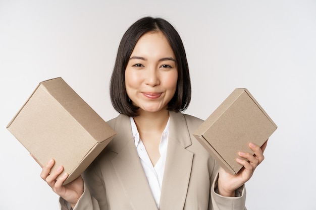 Immagine di commessa donna d'affari asiatica in possesso di scatole con prodotto di marca aziendale sorridente alla telecamera in piedi su sfondo bianco