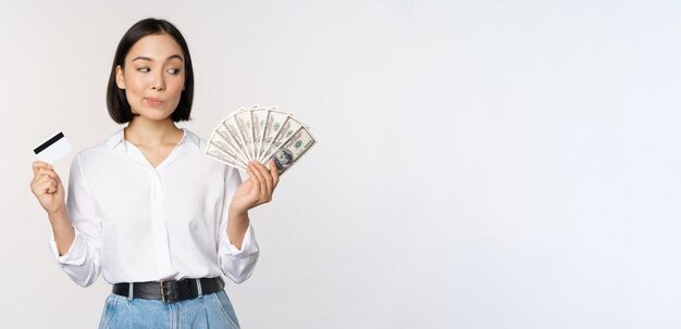 Immagine di donna asiatica guardando soldi dollari in possesso di carta di credito in un'altra mano pensando in piedi su sfondo bianco