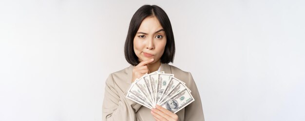 Immagine di donna d'affari asiatica felice imprenditrice mostrando denaro contanti dollari e pensando in piedi in tuta su sfondo bianco