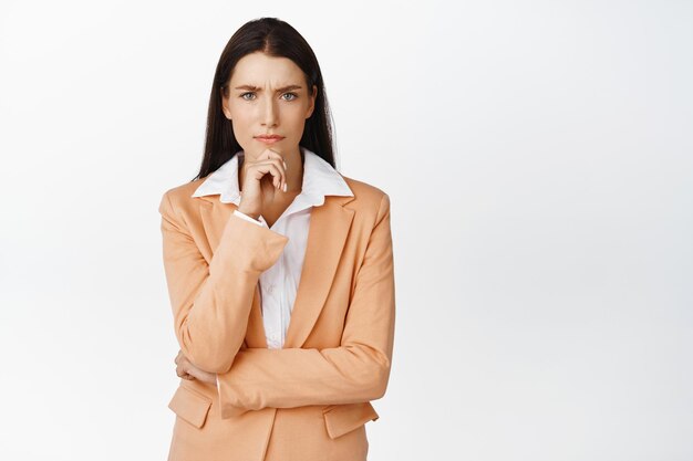 Immagine di donna d'affari che sembra preoccupata pensando con la fronte turbata accigliata che risolve il problema in piedi in tuta su sfondo bianco