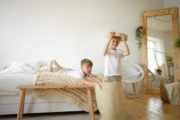Immagine di due adorabili scolari caucasici che si divertono in casa, giocano insieme nella camera dei genitori, sentendosi felici e spensierati. Bambini maschi carini che si divertono a casa