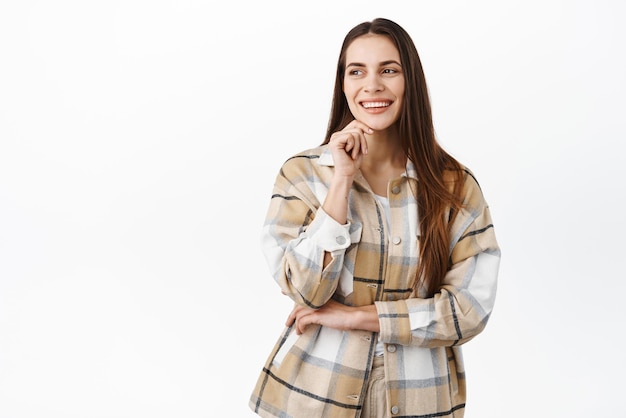 Immagine di elegante giovane donna professionale che tocca il viso sorridente e ridente sicura di sé guardando da parte il testo promozionale dello spazio di copia sulla sinistra in piedi su sfondo bianco