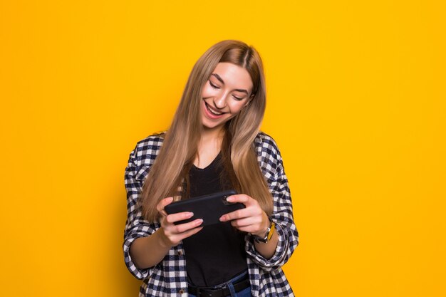 Immagine di felice carino bella giovane donna giocare con il telefono cellulare isolato sopra la parete gialla. Guardando da parte.