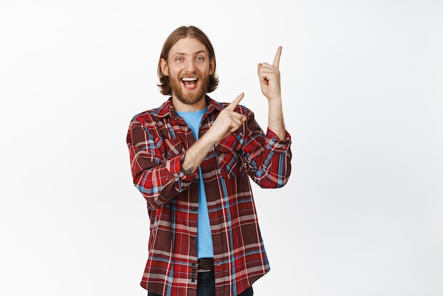 Immagine di felice hipster ragazzo biondo uomo barbuto che mostra banner fare clic sul collegamento puntando il dito nell'angolo in alto a destra e sorridente eccitato in piedi su sfondo bianco