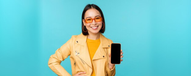 Immagine di giovane modello femminile asiatico alla moda in occhiali da sole alla moda che mostra l'applicazione dello schermo del telefono cellulare sull'interfaccia dello smartphone in piedi su sfondo blu