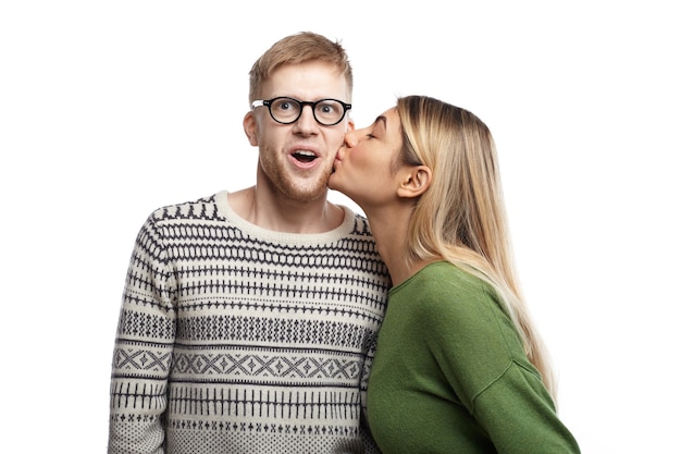 Immagine di giovane nerd divertente emotivo che indossa occhiali rettangolari esclamando eccitato, aprendo la bocca ampiamente come ragazza attraente che lo bacia sulla guancia. Persone, amore, romanticismo e appuntamenti