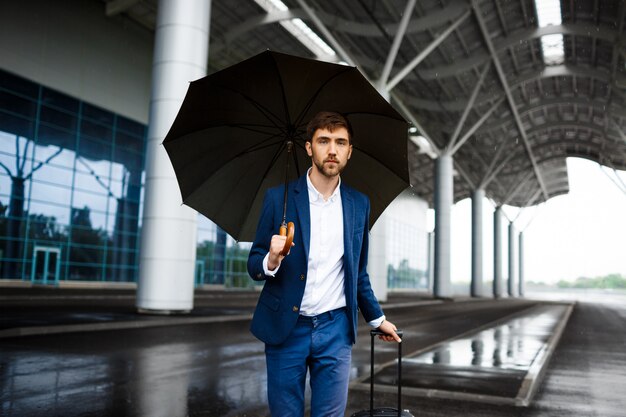 Immagine di giovane valigia della tenuta dell'uomo d'affari e dell'ombrello che stanno alla stazione piovosa