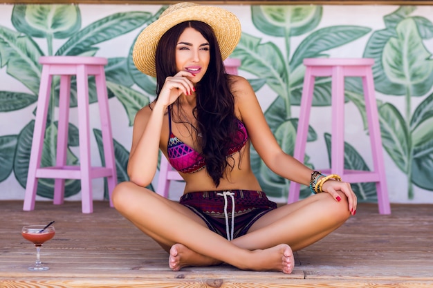 Immagine di moda estate di giovane donna in bikini sexy spiaggia con gustoso cocktail seduto sul pavimento di legno in elegante caffetteria