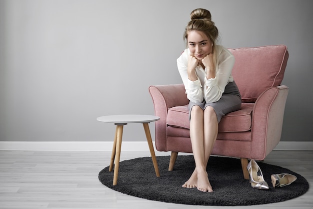 Immagine di positiva affascinante giovane imprenditrice europea che indossa eleganti abiti formali seduti in poltrona con i piedi nudi sul tappeto a casa, sorridendo misteriosamente, appoggiando il mento sulle sue mani