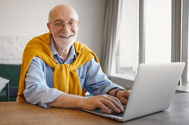 Immagine di successo positivo anziano barbuto blogger di viaggio europeo che scrive un articolo sul computer portatile, guardando e sorridendo che indossa un maglione elegante intorno al collo sopra la maglietta blu