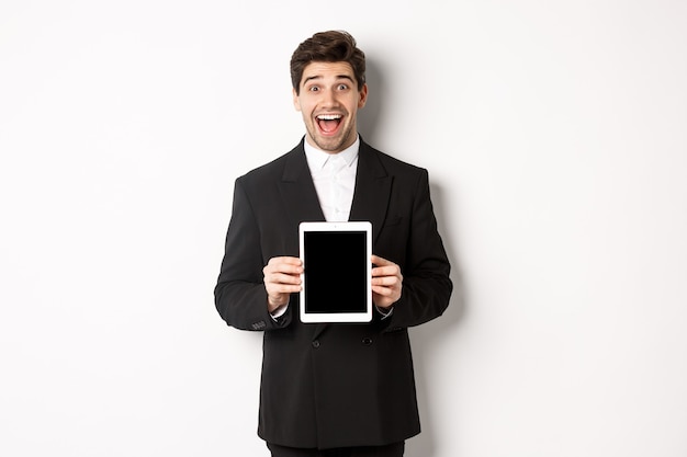 Immagine di un attraente imprenditore maschio in abito alla moda, che mostra lo schermo del tablet digitale e sorride stupito, in piedi su sfondo bianco.