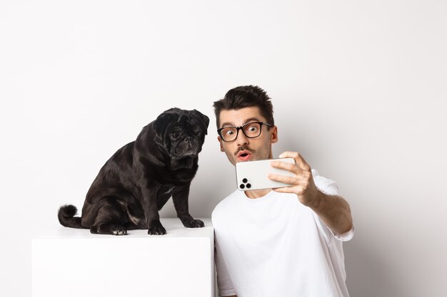 Immagine di un bel giovane che si fa selfie con un simpatico cane nero sullo smartphone, in posa con un carlino su sfondo bianco