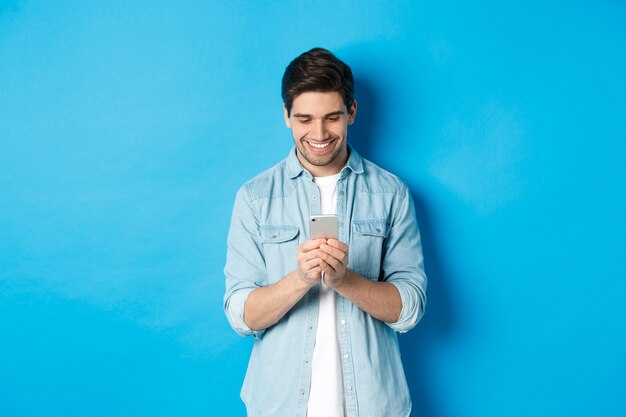 Immagine di un bel giovane che usa il telefono cellulare, manda un sms al telefono e sembra soddisfatto, in piedi su sfondo blu.