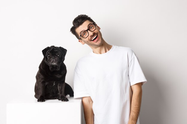 Immagine di un bel giovane in piedi vicino a un carlino nero carino e sorridente. Il proprietario del cane trascorre del tempo con il suo animale domestico, fissando la telecamera felice, sfondo bianco