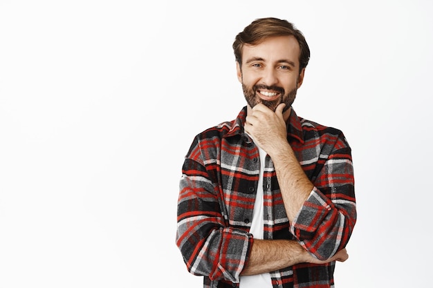 Immagine di un bel ragazzo contento che guarda smth interessante toccandosi la barba e pensando in piedi su sfondo bianco