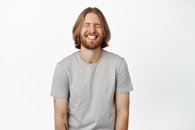 Immagine di un bel ragazzo spensierato, un uomo biondo che ride e sorride con gli occhi chiusi, in piedi in una maglietta grigia su sfondo bianco