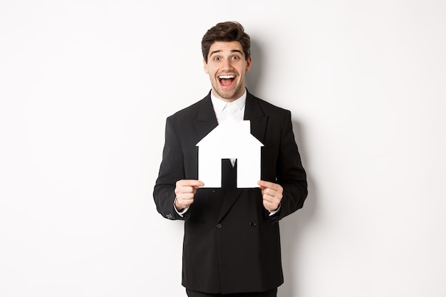 Immagine di un bell'agente immobiliare in abito nero che mostra il maket domestico e sembra stupito, vende case, in piedi su sfondo bianco