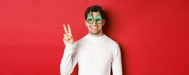 Immagine di un bell'uomo con un maglione bianco e occhiali da festa che mostra il segno della pace e sorride augurando buon...