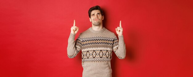 Immagine di un bell'uomo deluso e scettico che indossa un maglione natalizio che sorride e si acciglia mentre guarda...