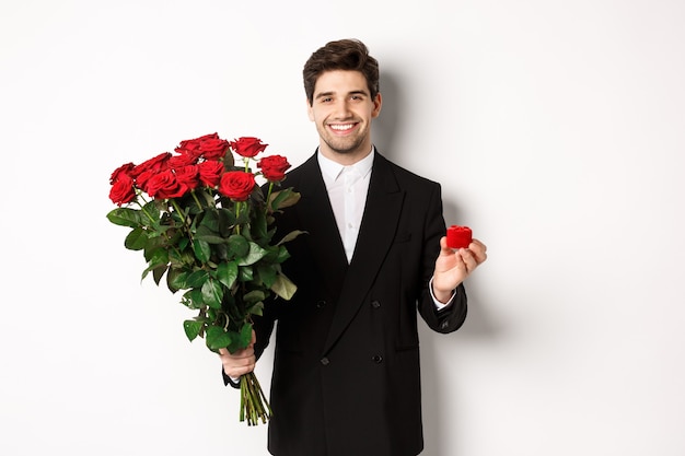 Immagine di un bell'uomo in abito nero, che tiene in mano un mazzo di rose rosse e un anello, fa una proposta, sorride fiducioso, in piedi su sfondo bianco.