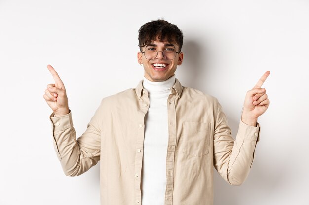 Immagine di un bell'uomo sorridente con gli occhiali che punta le dita lateralmente, mostrando pubblicità o varianti, in piedi allegro su sfondo bianco