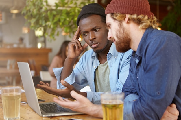 Immagine di un maschio afroamericano che utilizza un computer portatile durante l'incontro con il suo socio in affari caucasico al bar per discutere di strategia aziendale e piani su un paio di birre, persone e tecnologia