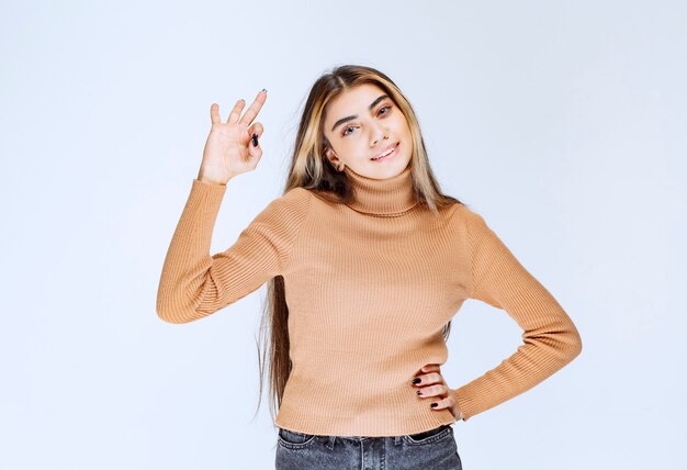 Immagine di un modello di giovane donna in maglione marrone in piedi e che mostra gesto ok.