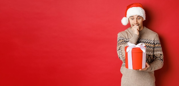 Immagine di un ragazzo preoccupato con cappello da Babbo Natale e maglione che guarda indeciso al regalo di Natale in piedi contro ...