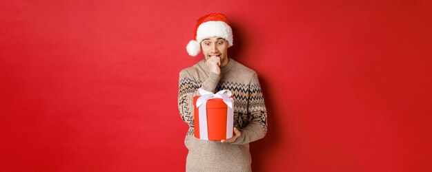 Immagine di un ragazzo preoccupato con cappello da Babbo Natale e maglione che sembra indeciso sul regalo di Natale in piedi contro