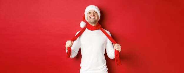 Immagine di un uomo angosciato e frustrato con il cappello da Babbo Natale che si soffoca con la sciarpa per la tristezza in piedi...