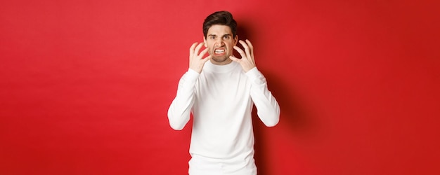 Immagine di un uomo arrabbiato e incazzato con un maglione bianco, che fa una smorfia e trema per la rabbia, in piedi furioso su sfondo rosso.