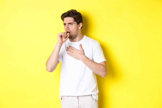 Immagine di un uomo con covid-19 o sintomi influenzali, tosse e sensazione di malessere, in piedi su sfondo giallo. Copia spazio