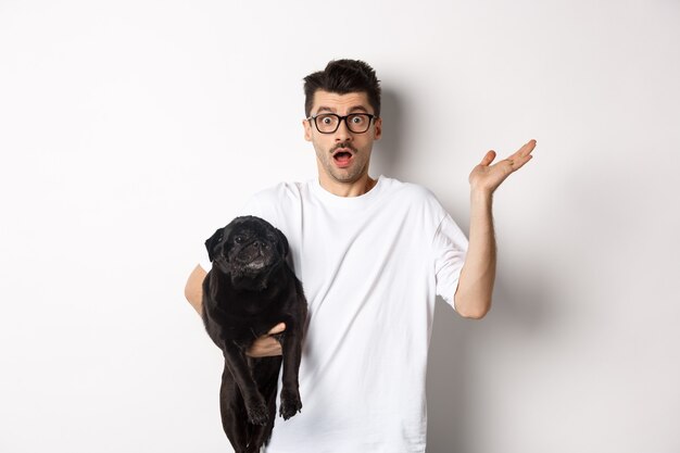 Immagine di un uomo confuso hipster che tiene il cane e alza le spalle, non so, alzando la mano perplesso, in piedi con il suo animale su sfondo bianco