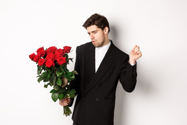 Immagine di un uomo elegante e sfacciato in abito nero, che sembra sicuro di sé e tiene in mano un mazzo di rose rosse, che va ad un appuntamento romantico, in piedi su sfondo bianco.